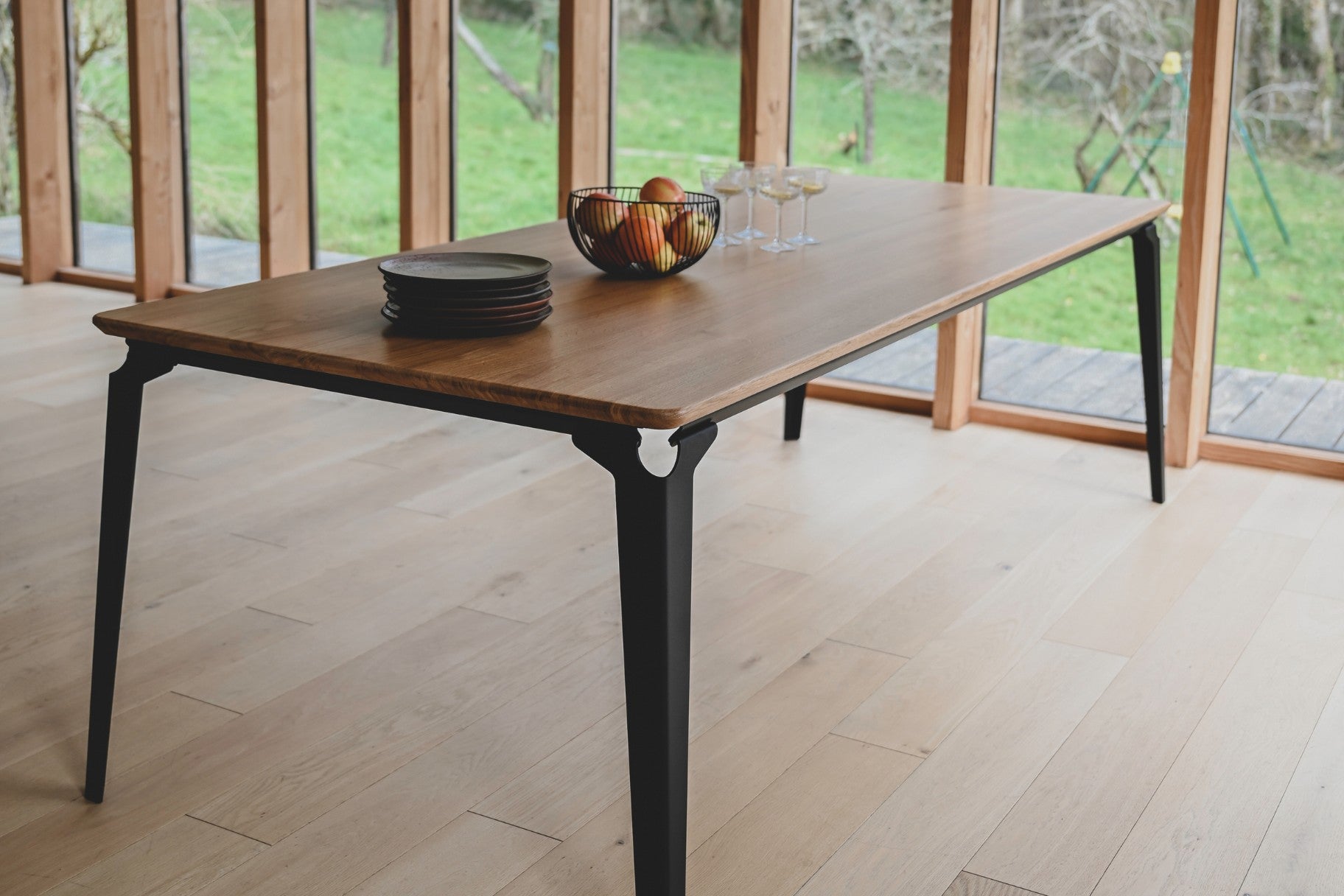 Table à Manger Angers en bois et acier noir avec une corbeille de fruits et des assiettes empilées.